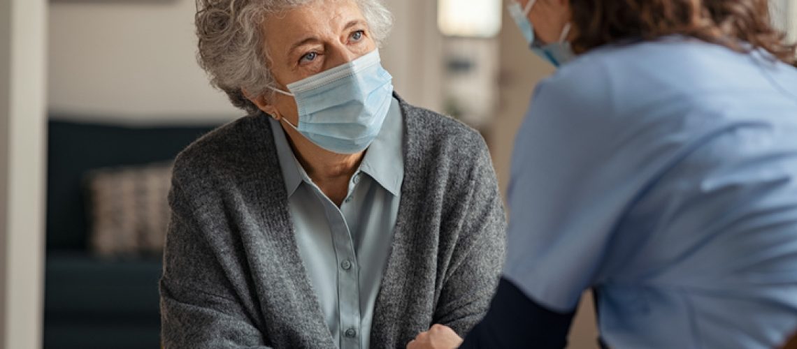 Elderly woman talking with a doctor while holding hands at home and wearing face protective mask. Worried senior woman talking to her general pratictioner visiting her at home during virus epidemic. Doctor explaining about precautionary measures during virus pandemic to old lady and takes care of her.