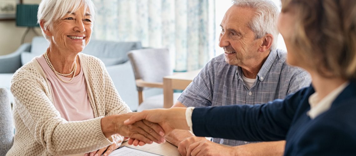 Happy old woman shaking hands with medical adviser for health insurance while sitting at home. Senior couple signing deal for insurance with financial advisor after retirement at home. Cheerful couple sealing with handshake a contract with financial advisor for investment.