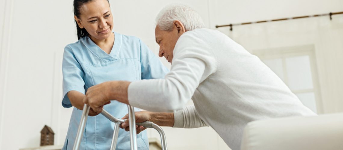 Professional caregiver. Joyful nice pleasant woman smiling and looking at the elderly man while helping him to walk