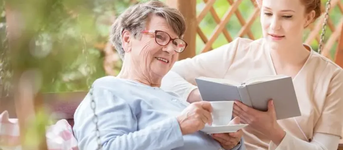 Happy elderly woman drinking tea while nurse reading book in the garden